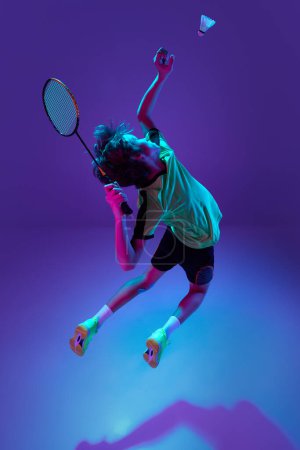Téléchargez les photos : Dynamic game. Teen boy in uniform playing badminton, hitting shuttlecock in a jump over blue purple background in neon ligth. Concept of sportive lifestyle, motion, action, competition - en image libre de droit