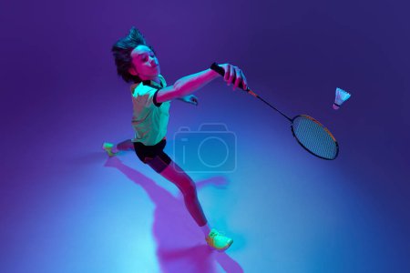 Téléchargez les photos : Top view. Portrait of teen boy in uniform playing badminton over blue purple background in neon ligth. Concept of sportive lifestyle, motion, action, competition, hobby - en image libre de droit