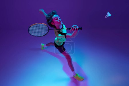 Téléchargez les photos : Top view. Portrait of teen boy in uniform playing badminton, returning shuttlecock in a run over blue purple background in neon ligth. Concept of sportive lifestyle, motion, action, competition - en image libre de droit