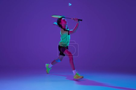 Téléchargez les photos : Dynamics. Portrait of teen boy in uniform playing badminton, hitting shuttlecock in a run over blue purple background in neon ligth. Concept of sportive lifestyle, motion, action, competition - en image libre de droit