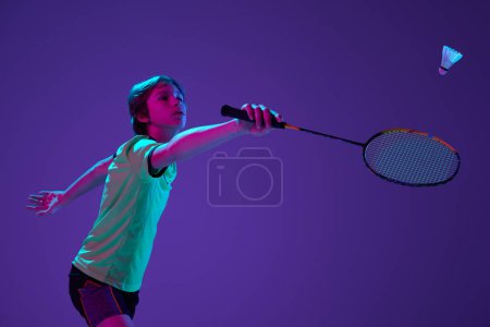 Téléchargez les photos : Portrait of teen boy in uniform playing badminton, hitting shuttlecock with racket isolated over blue purple background in neon ligth. Concept of sportive lifestyle, motion, action, competition - en image libre de droit