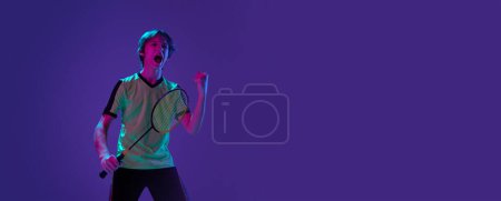 Téléchargez les photos : Portrait of teen boy in uniform, badminton player after successful game over purple background in neon ligth. Winning emotions. Banner. Concept of sportive lifestyle, motion, action, competition - en image libre de droit