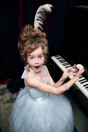 Téléchargez les photos : Emotive little girl, child in image of medieval royal person in fabulous dress sitting at the piano with makeup thing. Concept of historical remake, comparison of eras, medieval fashion, childhood - en image libre de droit