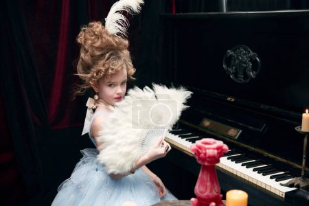 Téléchargez les photos : Cute little girl, child in image of medieval royal person in fabulous dress sitting at the piano with feather fan. Concept of historical remake, comparison of eras, medieval fashion, emotions - en image libre de droit