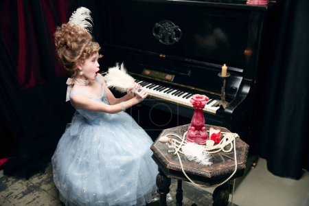 Foto de Cute little girl, child in image of medieval princess in fabulous dress sitting at the piano and emotionally singing in feather fan. Concept of historical remake, comparison of eras, medieval fashion - Imagen libre de derechos