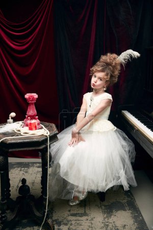 Téléchargez les photos : Cute little girl, child in image of medieval royal person in fabulous dress sitting at the piano and posing. Tenderness. Concept of historical remake, comparison of eras, medieval fashion, childhood - en image libre de droit