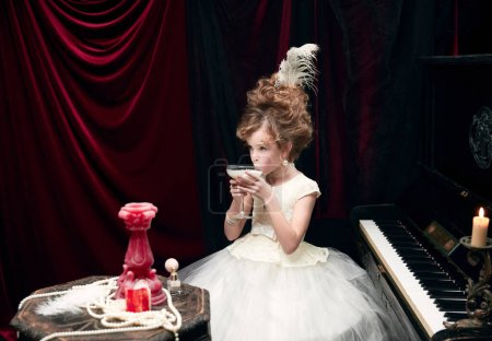 Téléchargez les photos : Cute little girl, child in image of medieval royal person sitting at the piano and drinking milkshake. Concept of historical remake, comparison of eras, medieval fashion, emotions, childhood - en image libre de droit