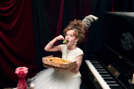 Téléchargez les photos : Portrait of little girl, child in image of medieval royal person in fabulous dress sitting at the piano and eating pizza. Concept of historical remake, comparison of eras, medieval fashion, emotions - en image libre de droit