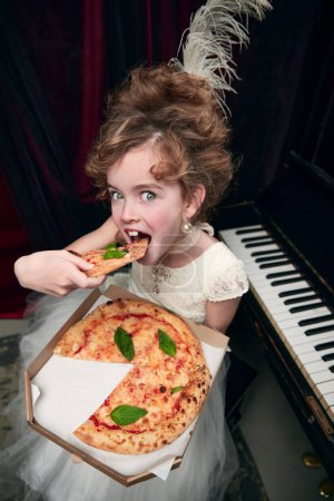 Téléchargez les photos : Portrait of little girl, child in image of medieval royal person sitting at the piano and eating pizza. Yummy. Concept of historical remake, comparison of eras, medieval fashion, emotions, childhood - en image libre de droit