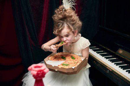 Téléchargez les photos : Portrait of cute little girl, child in image of medieval royal person sitting at the piano and emotionally eating pizza. Concept of historical remake, comparison of eras, medieval fashion, childhood - en image libre de droit