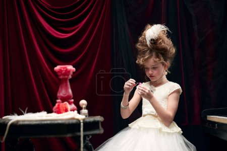 Téléchargez les photos : Portrait of cute little girl, child in image of medieval royal princess in white dress sitting at the piano and painting nails. Concept of historical remake, comparison of eras, fashion, childhood - en image libre de droit