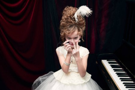 Foto de Portrait of cute little girl, child in image of medieval royal princess in white dress sitting at the piano and painting nails. Concept of historical remake, comparison of eras, fashion, childhood - Imagen libre de derechos