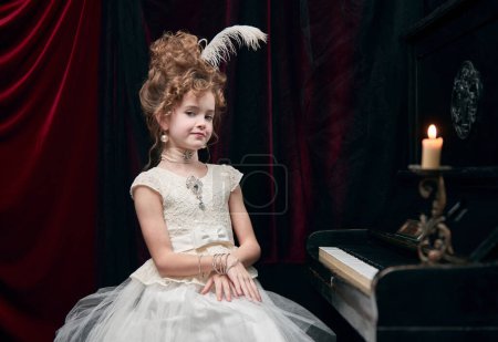 Cute little girl, child in image of medieval royal person, princess in fabulous dress sitting at the piano. Concept of historical remake, comparison of eras, medieval fashion, emotions, childhood