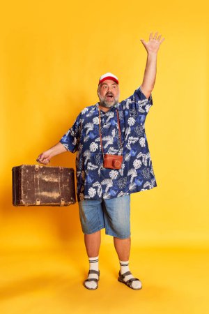 Téléchargez les photos : Portrait of emotive mature man in stylish shirt, cap posing with camera and suitcase over bright yellow background. Travel. Concept of american style, culture, emotions, facial expression, lifestyle - en image libre de droit