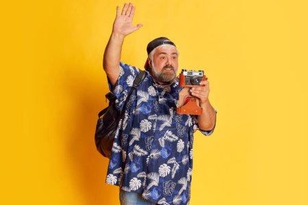 Téléchargez les photos : Portrait of fat mature man in stylish shirt posing, taking photo with vintage camera over bright yellow studio background. Concept of american style, culture, emotions, facial expression, travel - en image libre de droit
