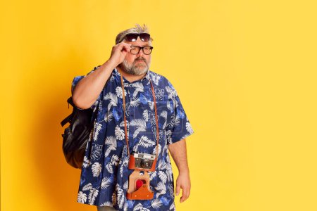 Photo for Portrait of mature man in stylish shirt, backpack, in glasses attentively looking, posing over bright yellow studio background. Concept of american style, culture, emotions, facial expression, travel - Royalty Free Image