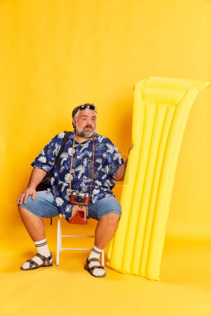 Téléchargez les photos : Portrait of fat mature man in stylish shirt sitting with swimming mattress, posing over bright yellow studio background. Concept of american style, culture, emotions, facial expression, travel - en image libre de droit