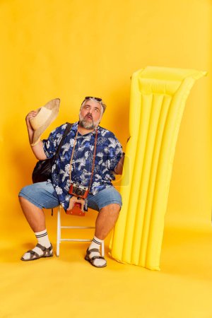 Photo for Portrait of fat mature man in stylish shirt sitting with swimming mattress, posing over bright yellow background. Summer, hot. Concept of american style, culture, emotions, facial expression, travel - Royalty Free Image