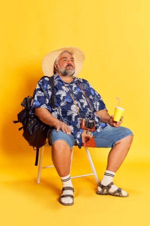 Téléchargez les photos : Portrait of fat mature man in stylish shirt, hat and camera posing over bright yellow studio background. Traveling look. Concept of american style, culture, emotions, facial expression, travel - en image libre de droit