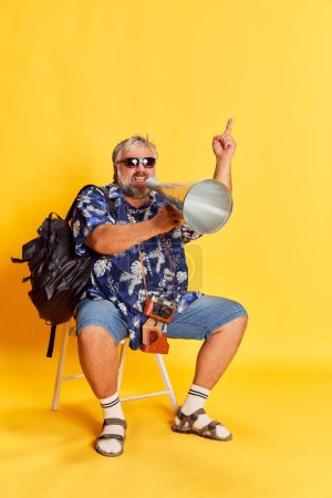 Photo for Portrait of fat mature man posing, sitting and shouting in megaphone over bright yellow studio background. Information. Concept of american style, culture, emotions, facial expression, travel - Royalty Free Image