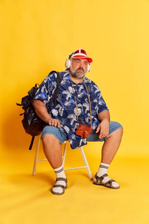 Téléchargez les photos : Portrait of fat mature man in stylish shirt, cap, with backpack sitting with bored face, posing over bright yellow background. Concept of american style, culture, emotions, facial expression, travel - en image libre de droit