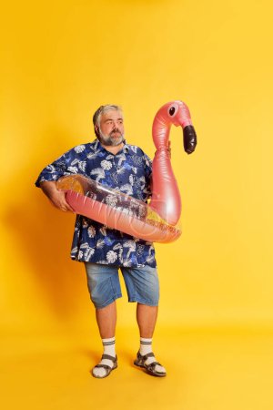 Foto de Portrait of fat mature man in stylish shirt posing in pink swimming circle on bright yellow studio background. Summer vacation. Concept of american style, culture, emotions, facial expression, travel - Imagen libre de derechos