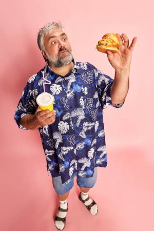 Foto de Portrait of emotive mature man in colorful shirt posing with delicious burger over pink studio background. Fast food taste. Concept of american style, culture, emotions, facial expression, lifestyle - Imagen libre de derechos