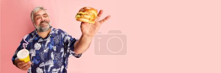 Photo for Portrait of mature man in colorful shirt smiling, posing with delicious burger on pink studio background. Fast food. Banner. Concept of american style, culture, emotions, facial expression, lifestyle - Royalty Free Image
