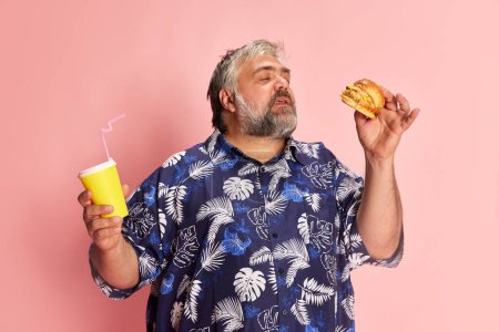 Téléchargez les photos : Portrait of mature man in colorful shirt posing with delicious burger over pink studio background. Favourite taste. Concept of american style, culture, emotions, facial expression, lifestyle - en image libre de droit
