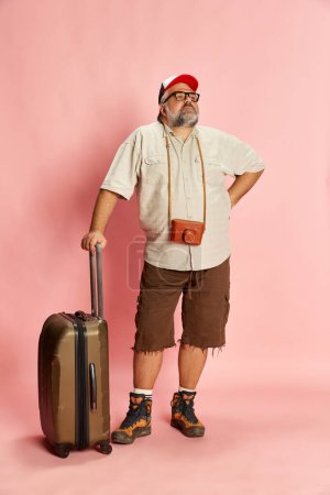 Foto de Portrait of mature overweight man in casual clothes with suitcase and vintage camera posing over pink background. Tourist. Concept of american style, culture, emotions, facial expression, lifestyle - Imagen libre de derechos