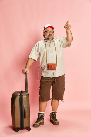 Foto de Portrait of mature overweight man, tourist in casual clothes with suitcase and vintage camera posing over pink background. Concept of american style, culture, emotions, facial expression, lifestyle - Imagen libre de derechos