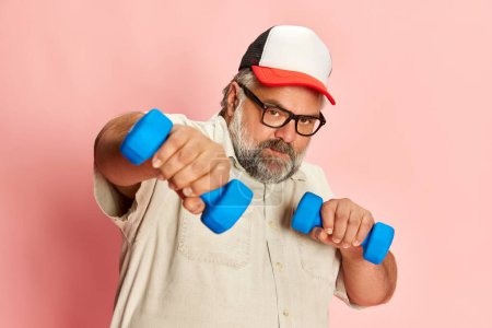 Foto de Portrait of mature overweight man training with dumbbells over pink studio background. Motivation to start sportive routine. Concept of american style, culture, emotions, facial expression, lifestyle - Imagen libre de derechos