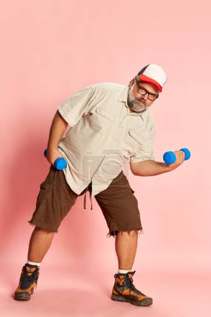 Téléchargez les photos : Portrait of mature overweight man training with dumbbells over pink studio background. Starting healthy lifestyle. Concept of american style, culture, emotions, facial expression, sport, fitness - en image libre de droit