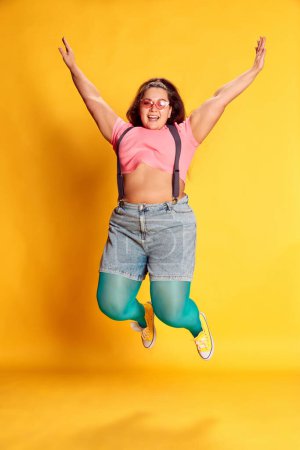 Téléchargez les photos : Young overweight woman in casual bright clothes posing, jumping on vivid yellow studio background. Happy and positive vibes. Concept of american style, culture, emotions, facial expression, lifestyle - en image libre de droit