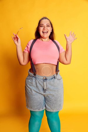 Téléchargez les photos : Super happy. Portrait of young overweight woman in casual bright clothes posing with smile on vivid yellow studio background. Concept of american style, culture, emotions, facial expression, lifestyle - en image libre de droit