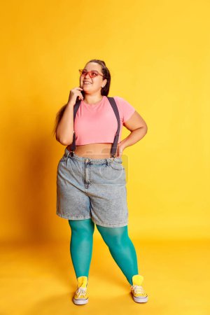Foto de Portrait of young overweight woman in casual bright clothes posing over vivid yellow studio background. Dreamy look. Concept of american style, culture, emotions, facial expression, lifestyle - Imagen libre de derechos