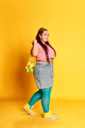 Foto de Portrait of young overweight woman posing with bag with various vegetables over vivid yellow background. Grocery shopping. Concept of american style, culture, emotions, facial expression, lifestyle - Imagen libre de derechos