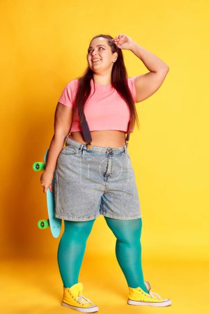 Foto de Portrait of young overweight woman in casual bright clothes posing with skateboard on vivid yellow background. Active hobby. Concept of american style, culture, emotions, facial expression, lifestyle - Imagen libre de derechos