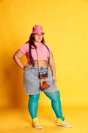 Téléchargez les photos : Portrait of young overweight woman, traveller in casual bright clothes posing with camera on vivid yellow studio background. Concept of american style, culture, emotions, facial expression, lifestyle - en image libre de droit