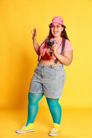 Téléchargez les photos : Portrait of young overweight woman, traveller in casual bright clothes, taking photo with camera over yellow background. Concept of american style, culture, emotions, facial expression, lifestyle - en image libre de droit