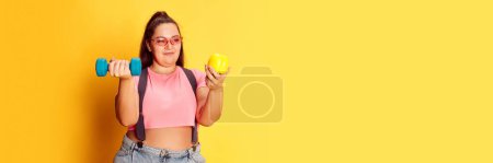 Photo for Portrait of overweight young woman posing with dumbbell and apple over yellow studio background. Diet and sport. Concept of american style, culture, emotions, facial expression, lifestyle. Banner - Royalty Free Image