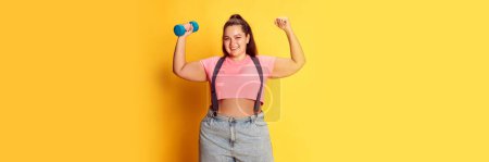Téléchargez les photos : Portrait of overweight young woman posing with dumbbells over yellow studio background. Maintaining healthy lifestyle. Concept of american style, culture, emotions, facial expression, sport, body care - en image libre de droit
