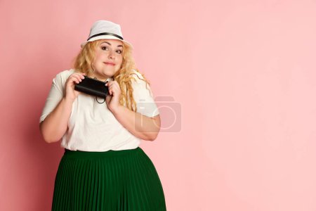 Téléchargez les photos : Portrait of stylish, beautiful woman posing in hat, white T-shirt and green skirt over pink studio background. Concept of american style, culture, emotions, facial expression, lifestyl, fashion - en image libre de droit
