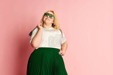 Foto de Portrait of stylish, beautiful woman posing in hat, white T-shirt and green skirt over pink studio background. Concept of american style, culture, emotions, facial expression, lifestyl, fashion - Imagen libre de derechos