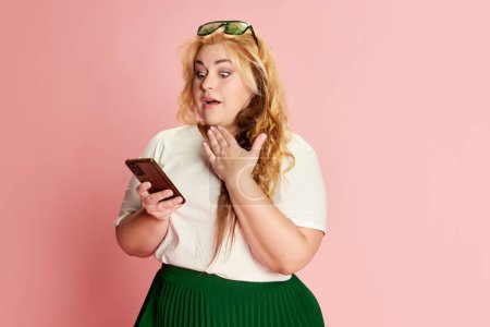 Foto de Portrait of stylish woman posing, emotionally looking on phone over pink background. Communication, reading messages, news. Concept of american style, culture, emotions, facial expression, lifestyle - Imagen libre de derechos