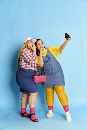 Photo for Portrait of two women, friends taking photo with camera, posing over blue studio background. Cheerful meeting. Concept of american style, culture, emotions, facial expression, lifestyle - Royalty Free Image