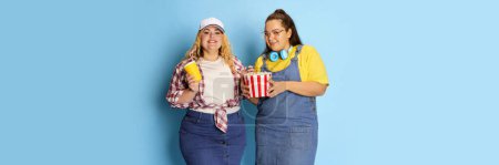 Foto de Portrait of two fat, beautiful women, friends posing with popcorn basket over blue studio background. Movie time. Banner. Concept of american style, culture, emotions, facial expression, lifestyle - Imagen libre de derechos