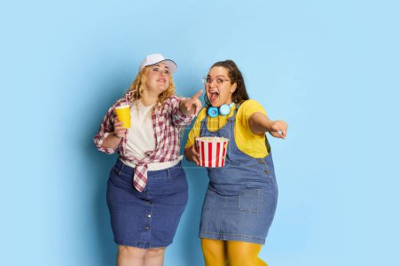 Foto de Portrait of two fat, beautiful women, friends posing with popcorn basket over blue studio background. Comedy movie. Concept of american style, culture, emotions, facial expression, lifestyle - Imagen libre de derechos