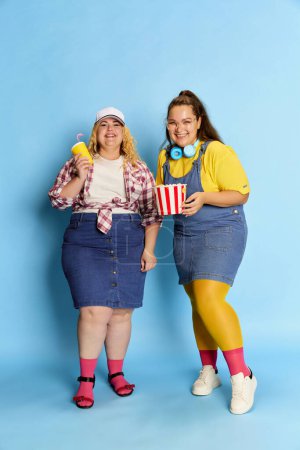 Photo for Portrait of two fat, beautiful women, friends posing with popcorn basket over blue studio background. Weekend leisure time. Concept of american style, culture, emotions, facial expression, lifestyle - Royalty Free Image
