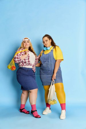 Photo for Portrait of two fat, beautiful women, friends posing with bag with fruits and vegetables over blue studio background. Diet. Concept of american style, culture, emotions, facial expression, lifestyle - Royalty Free Image
